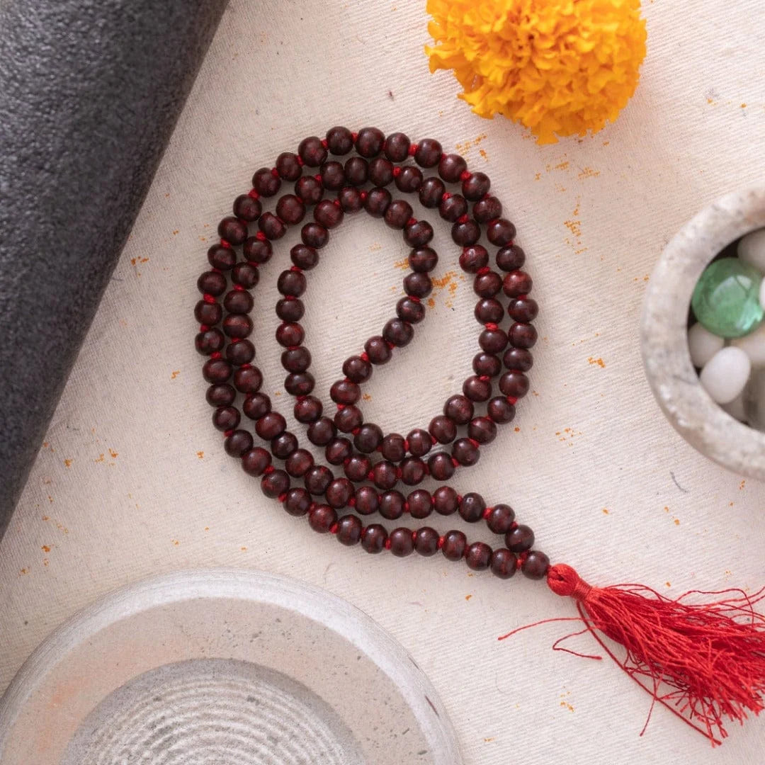 Brown Natural Tulsi Mala with 108 small beads, featuring a red tassel, displayed on a textured surface with decorative elemen